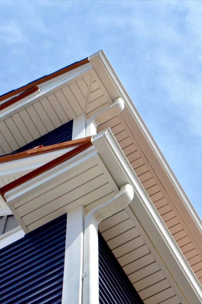 Corner of a house with white gutters and trim, beige siding, and dark blue panels against a clear blue sky.