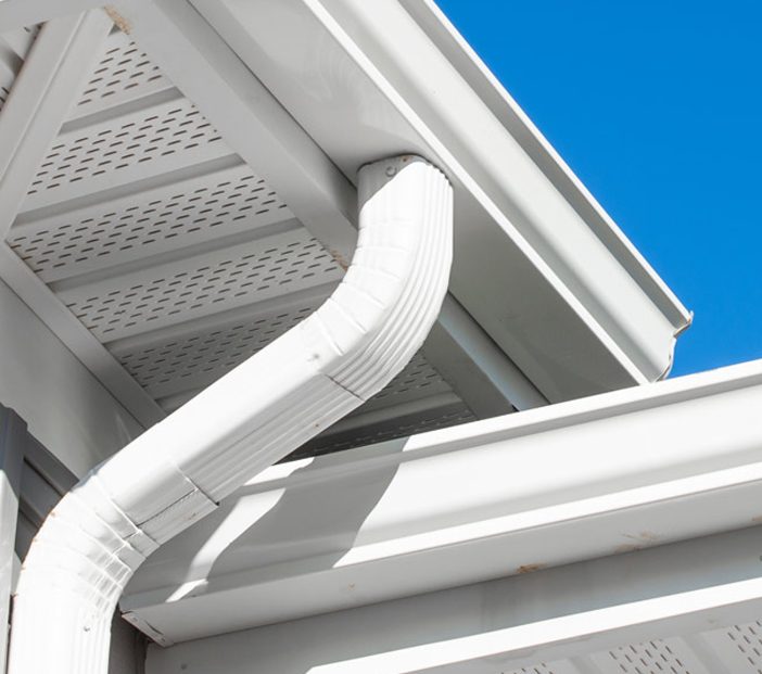 Close-up of a white rain gutter and downspout on a light-colored building with a blue sky backdrop.