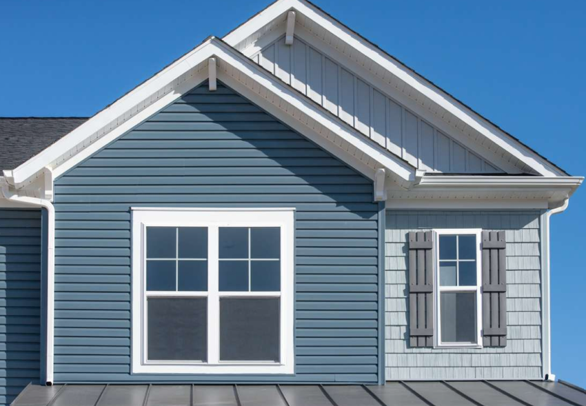 Blue siding house with white-framed window and brown shutters under a peaked roof against a clear sky.